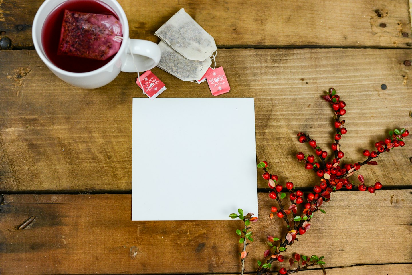 A white mug of tea sits on wood with berries and stationery.